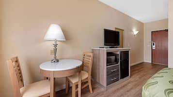 A hotel room with a round table, two wooden chairs, a television, a minibar, and a bed with a green patterned bedspread.