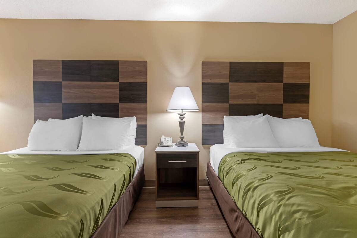A hotel room with two beds, each with green patterned bedding, a bedside table with a lamp, and a wall with a checkered wood panel design.