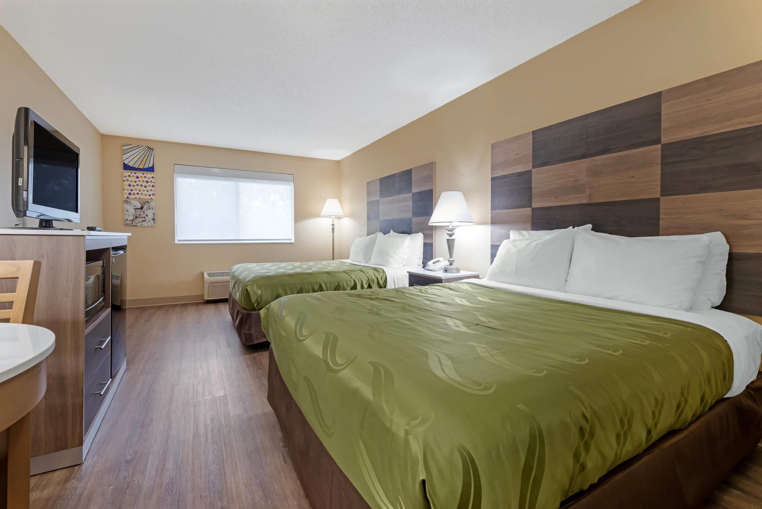 Habitación de hotel con dos camas, un escritorio con televisor, una ventana y una pared con estampado de cuadros.