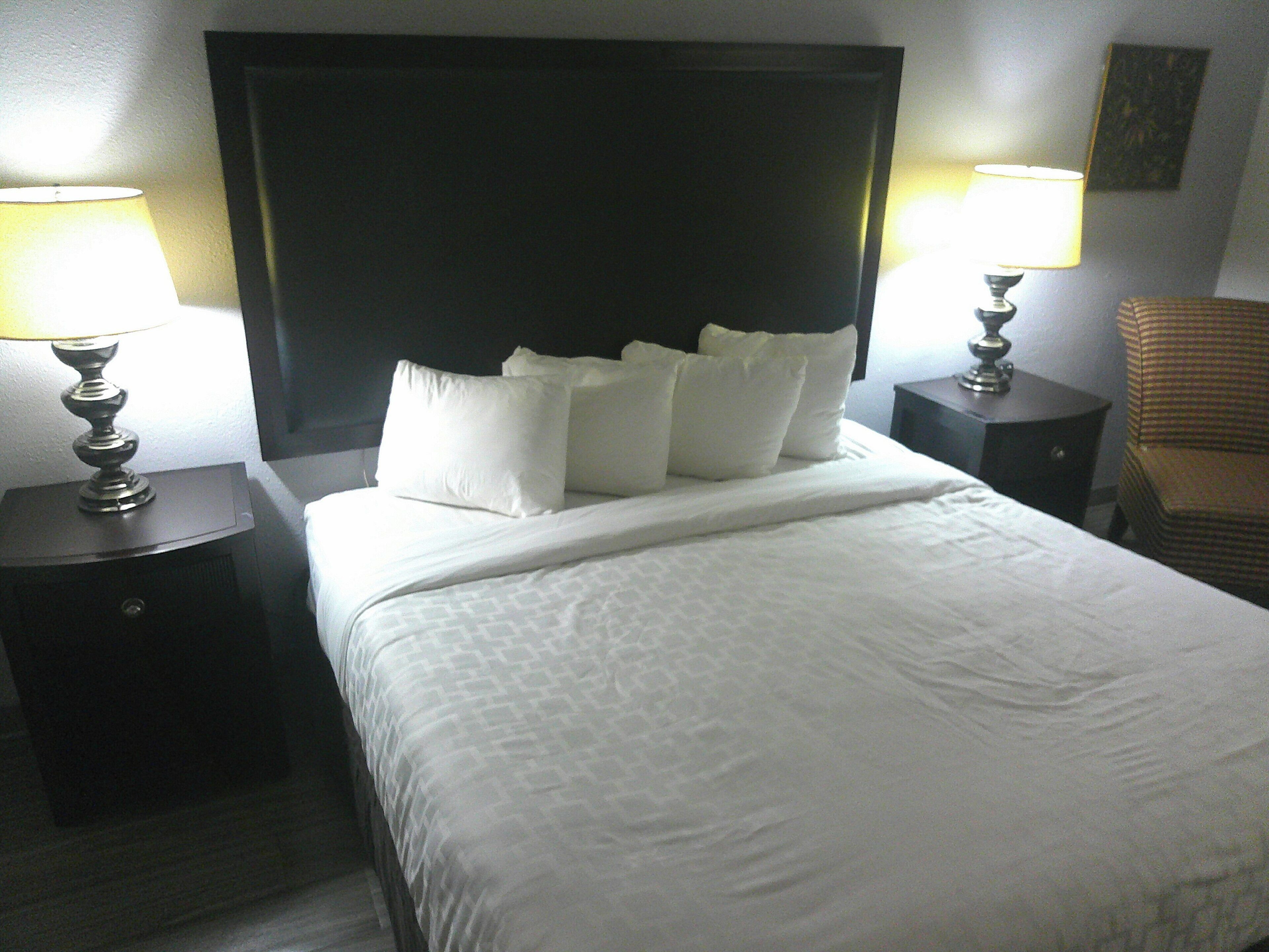 A neatly made bed with white linens and pillows, two bedside tables with lamps, and a chair in a hotel room.