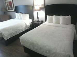Two single beds with white bedding, a dark headboard, a bedside table with a lamp, and a framed picture on the wall.