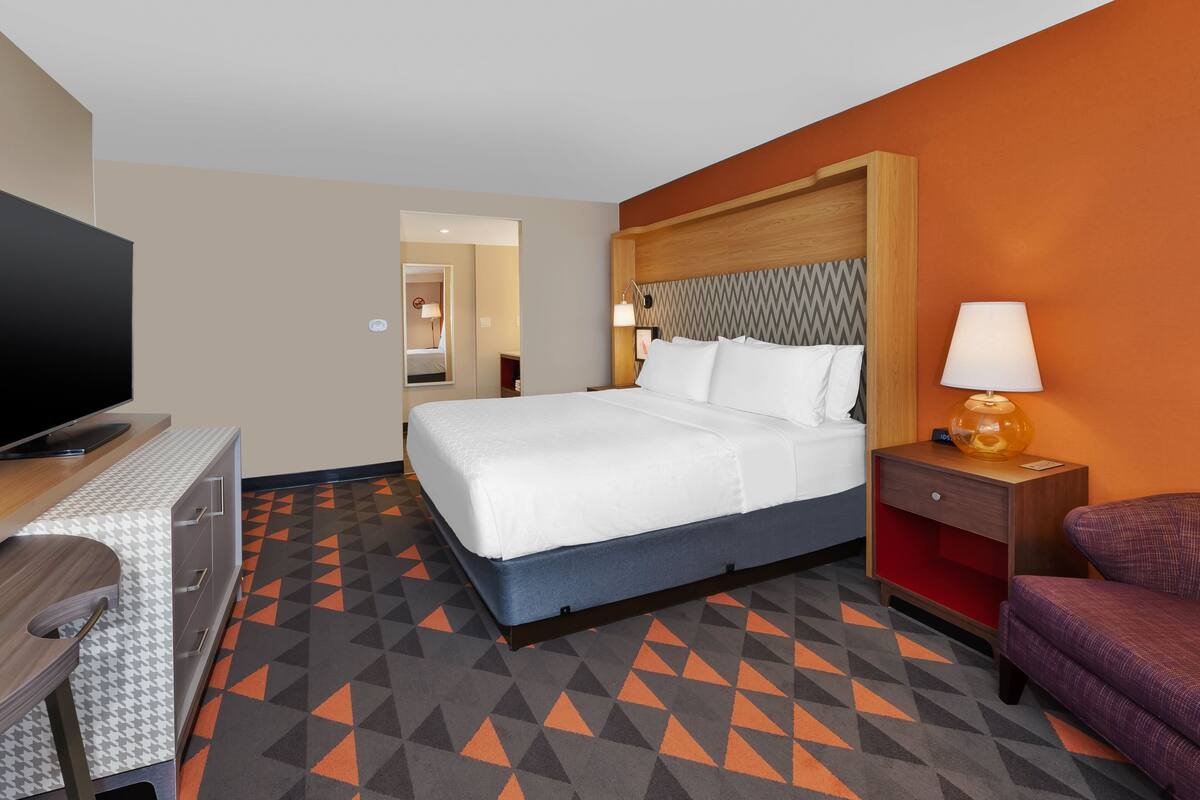 Une chambre d’hôtel comprenant un lit, un bureau avec une télévision, une chaise, une table de chevet avec une lampe et une moquette à motifs.