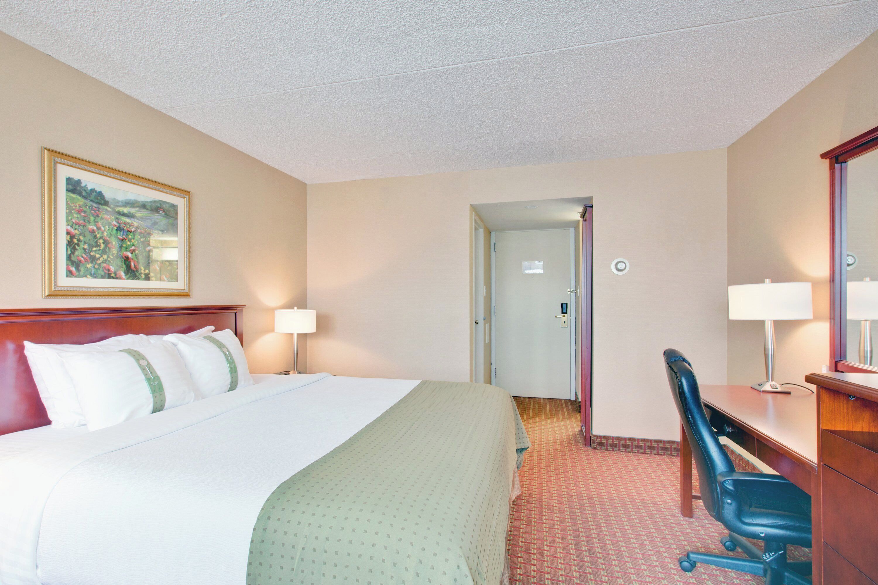 A hotel room with a bed, a desk, a chair, a desk lamp, a bedside table, a mirror, and a door leading to another room.