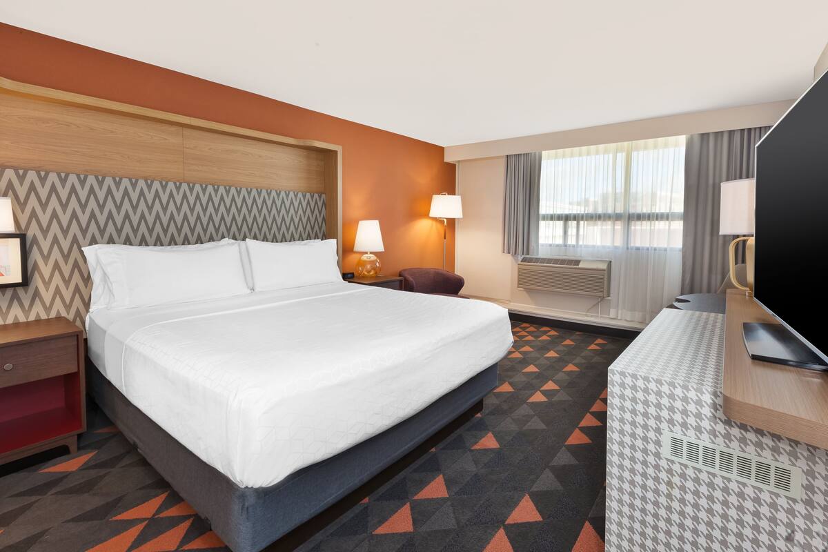 Une chambre d’hôtel avec un lit, des tables de chevet, un bureau avec une télévision et une fenêtre avec des rideaux.