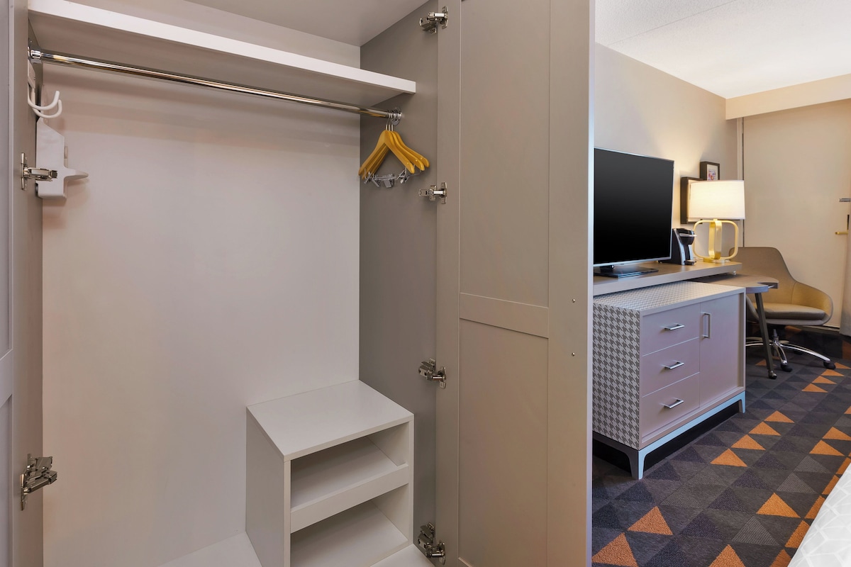 A hotel room with a wardrobe, a television, a desk with a lamp, and a patterned carpet.