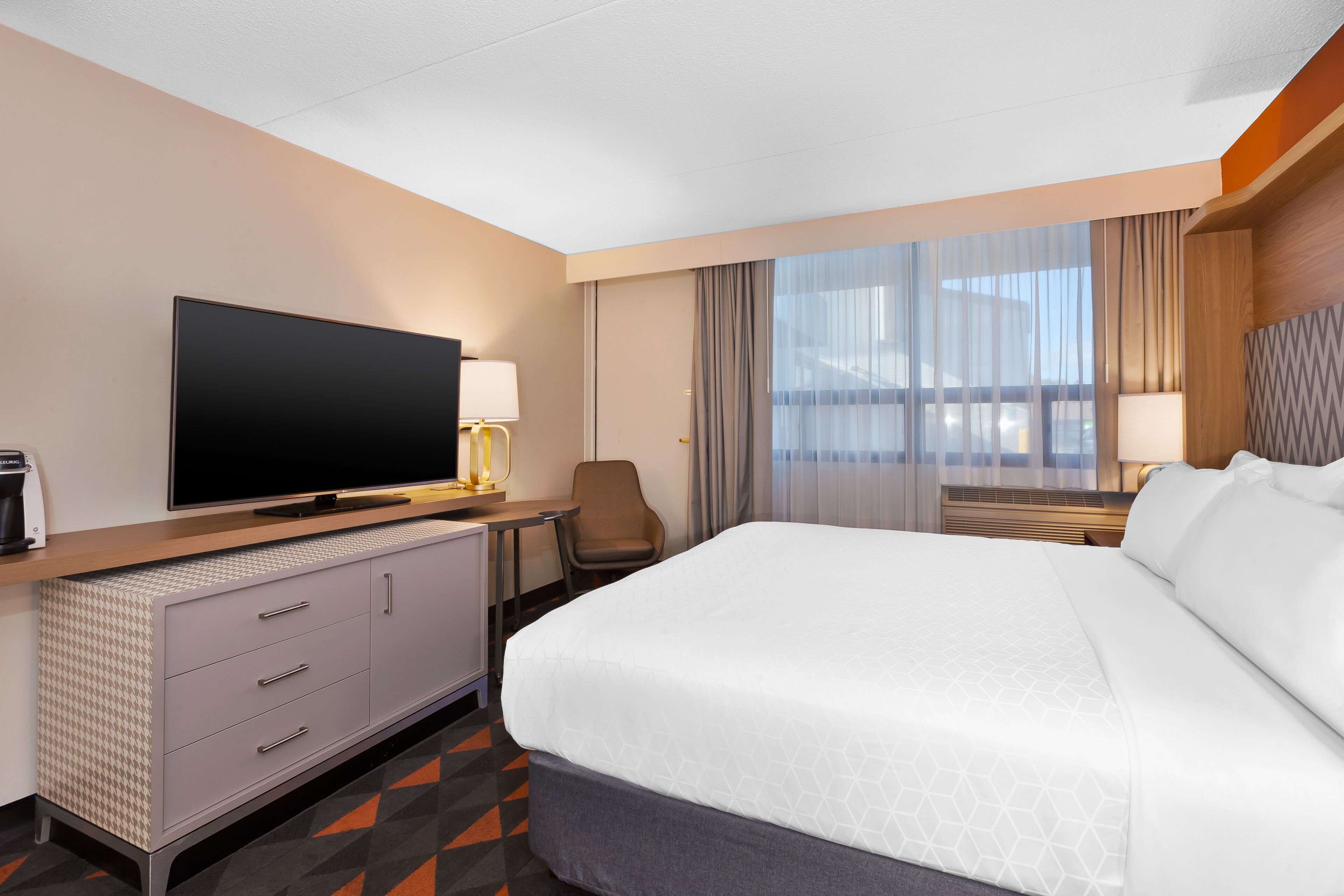 Une chambre d’hôtel dotée d’un grand lit, d’un téléviseur à écran plat, d’un bureau avec une lampe et offrant une vue sur la ville grâce à de grandes fenêtres.