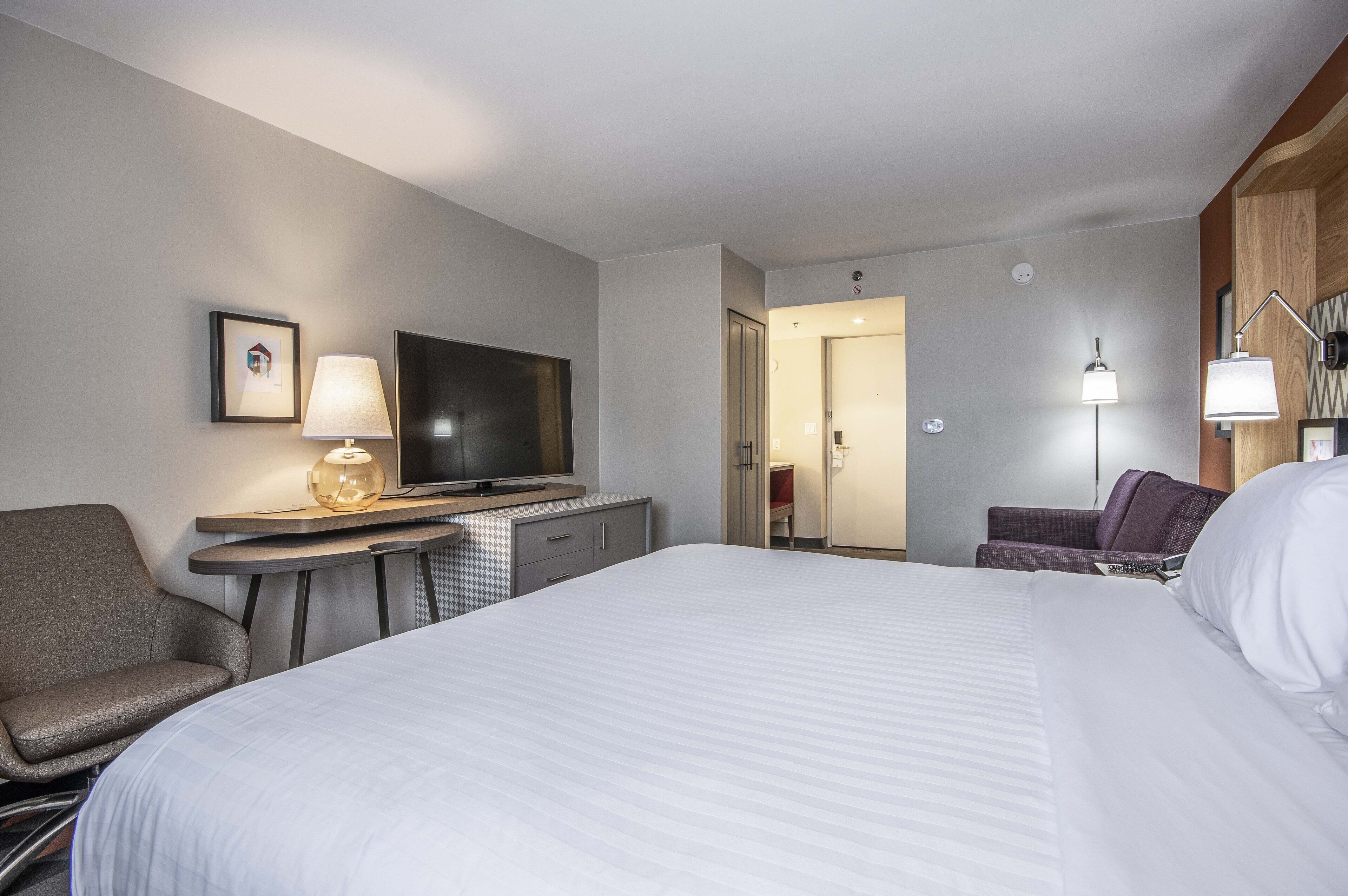 Una habitación de hotel con una cama grande, un escritorio con televisor, una silla y una lámpara.