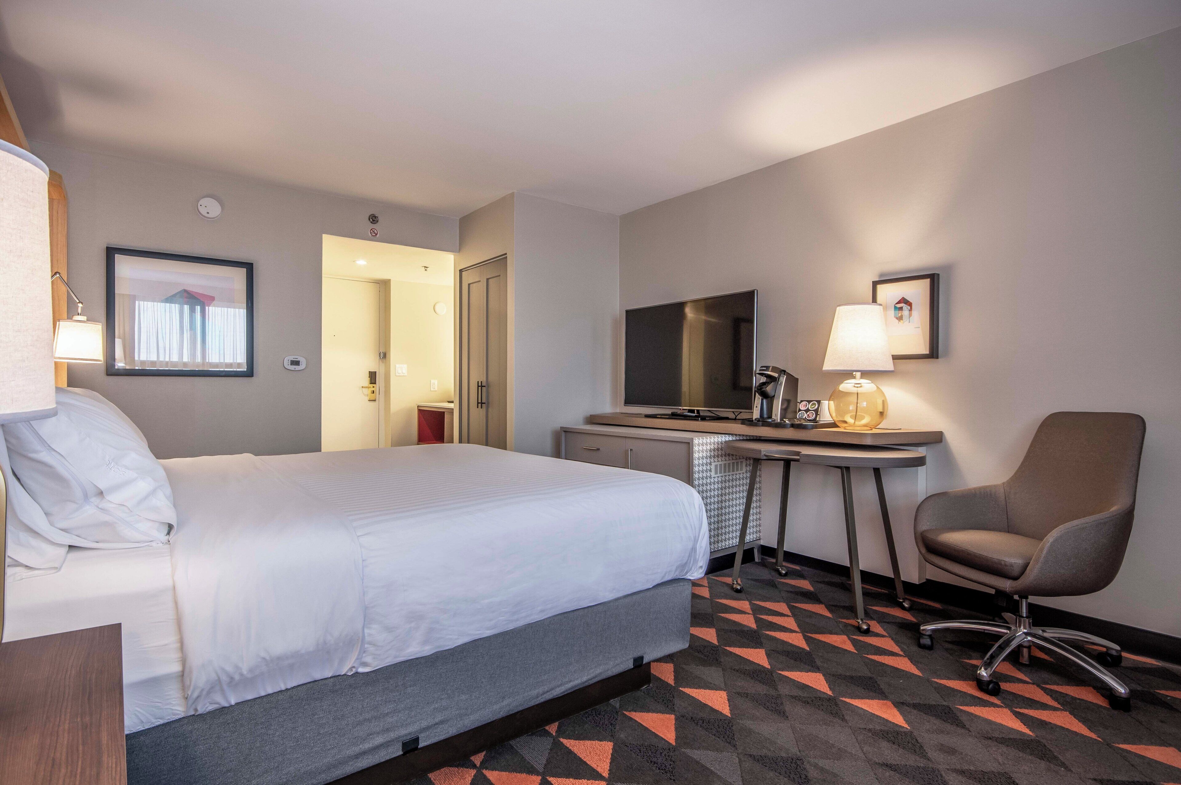 A hotel room with a bed, desk, chair, television, and a patterned carpet.