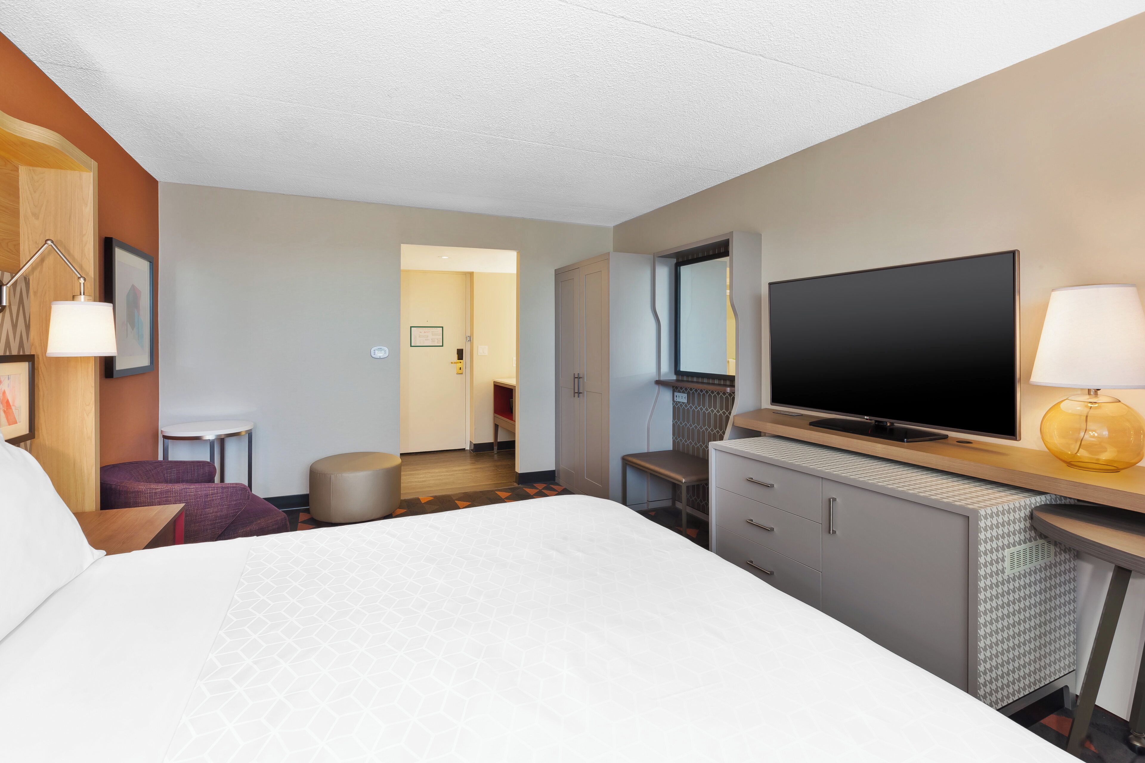 Quarto de hotel com uma cama grande, uma escrivaninha, televisão de tela plana, uma cadeira, uma mesinha e um abajur.