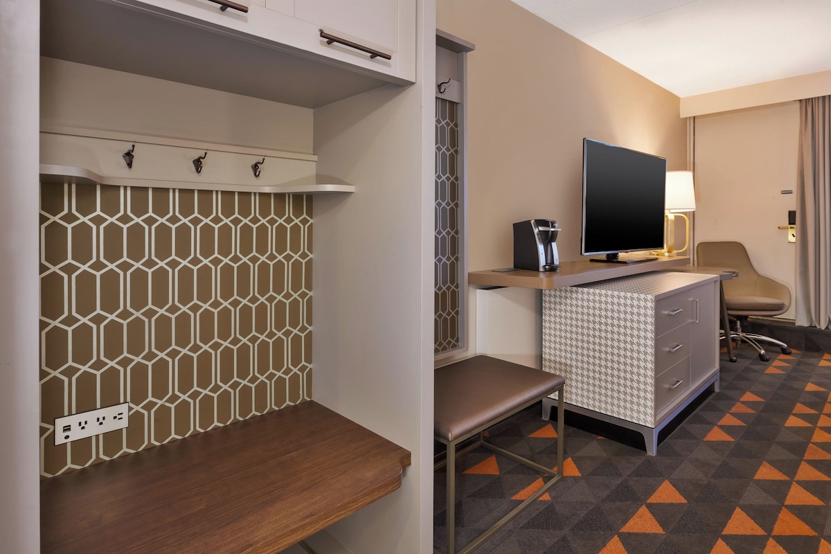 A hotel room with a patterned wall, a desk with a flat-screen TV, a lamp, a chair, and a patterned carpet.