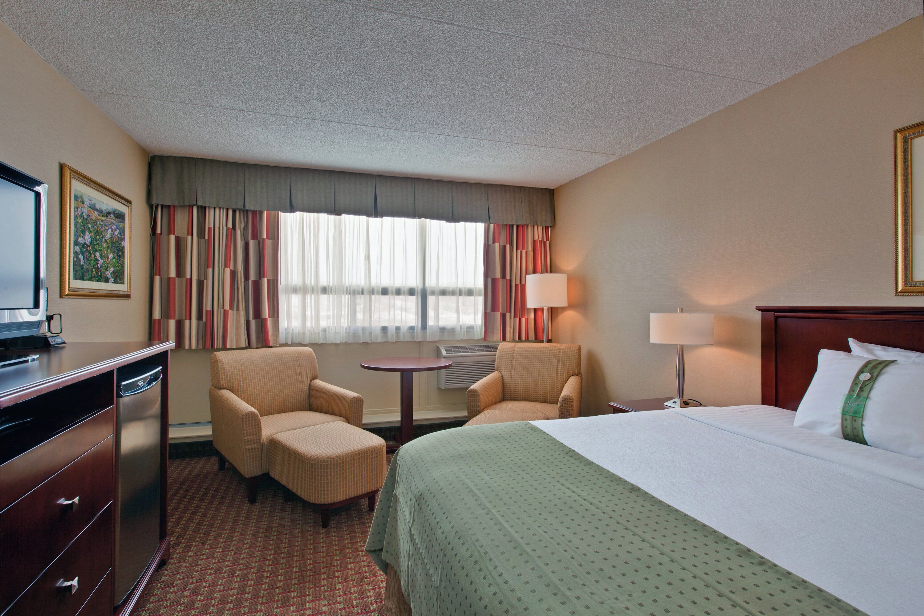 A hotel room with a bed, two armchairs, a desk with a television, a lamp, and a window with curtains.