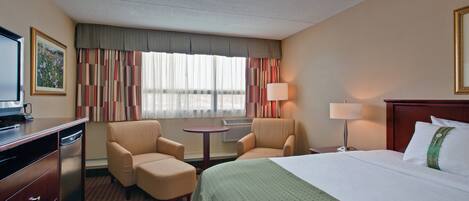 A hotel room with a bed, two armchairs, a desk with a television, a lamp, and a window with curtains.