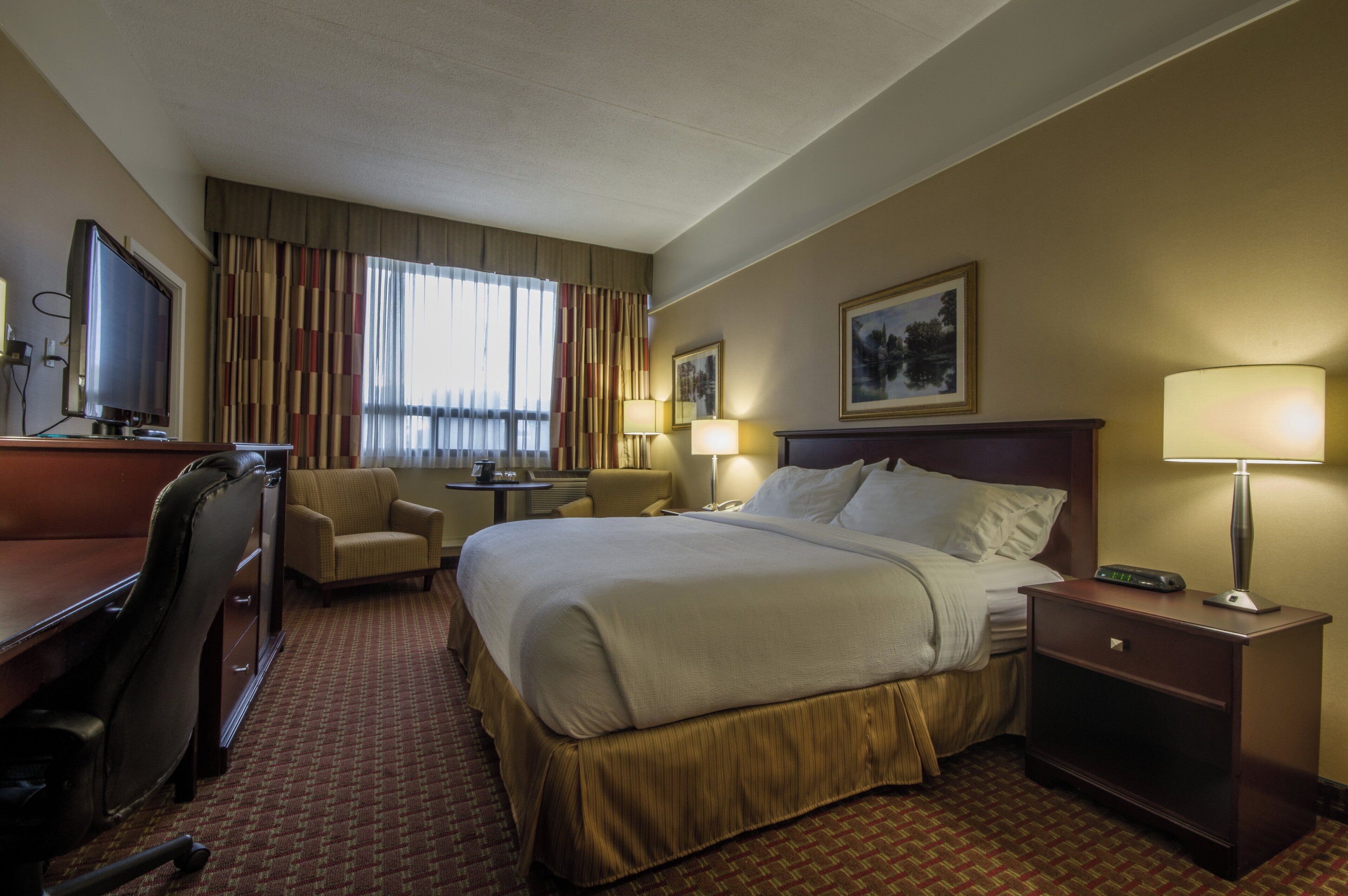 A hotel room with a bed, a desk, a chair, a TV, and a lamp.