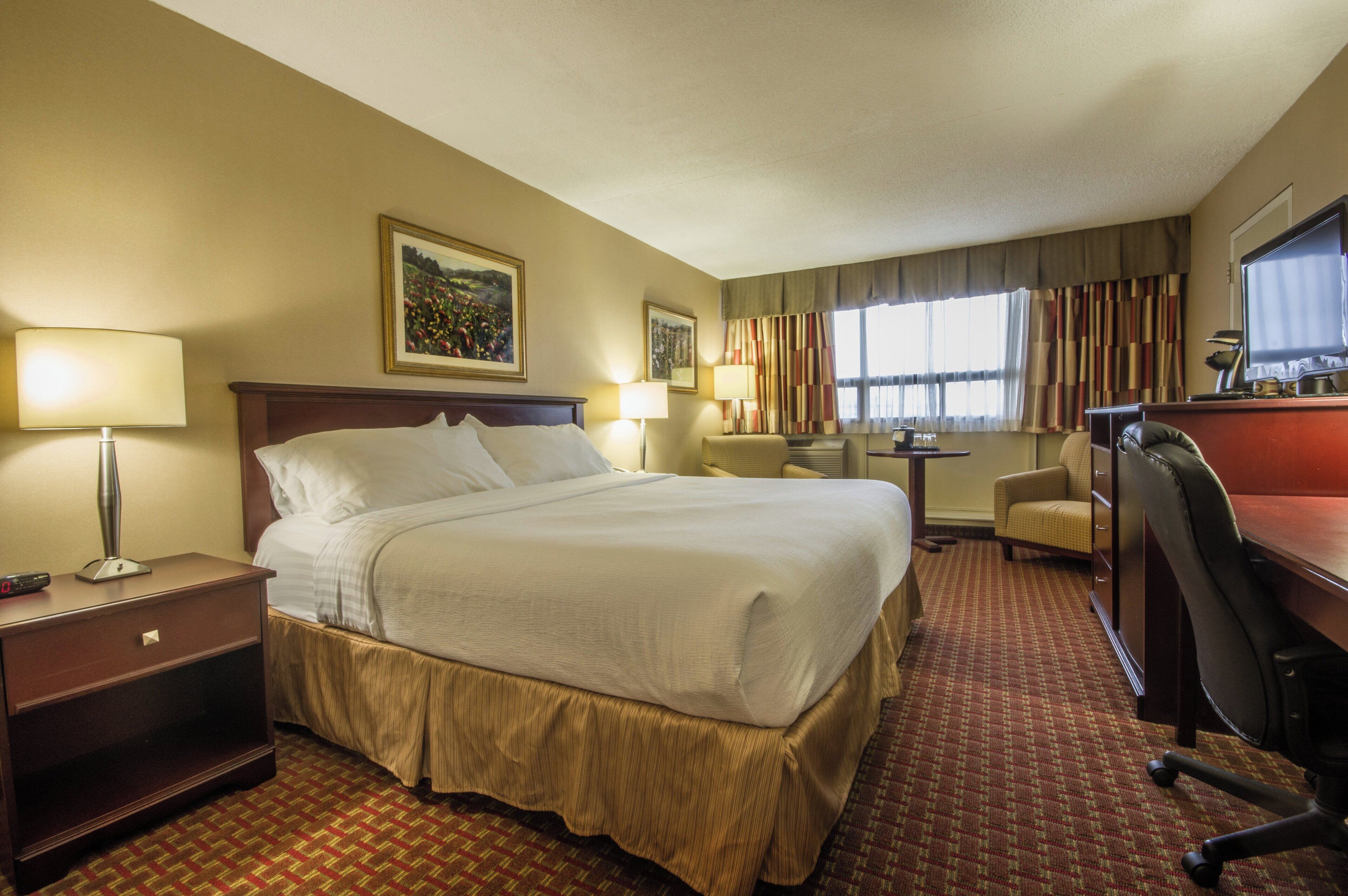 A hotel room with a bed, bedside lamps, a desk with a computer, a chair, and a window with curtains.