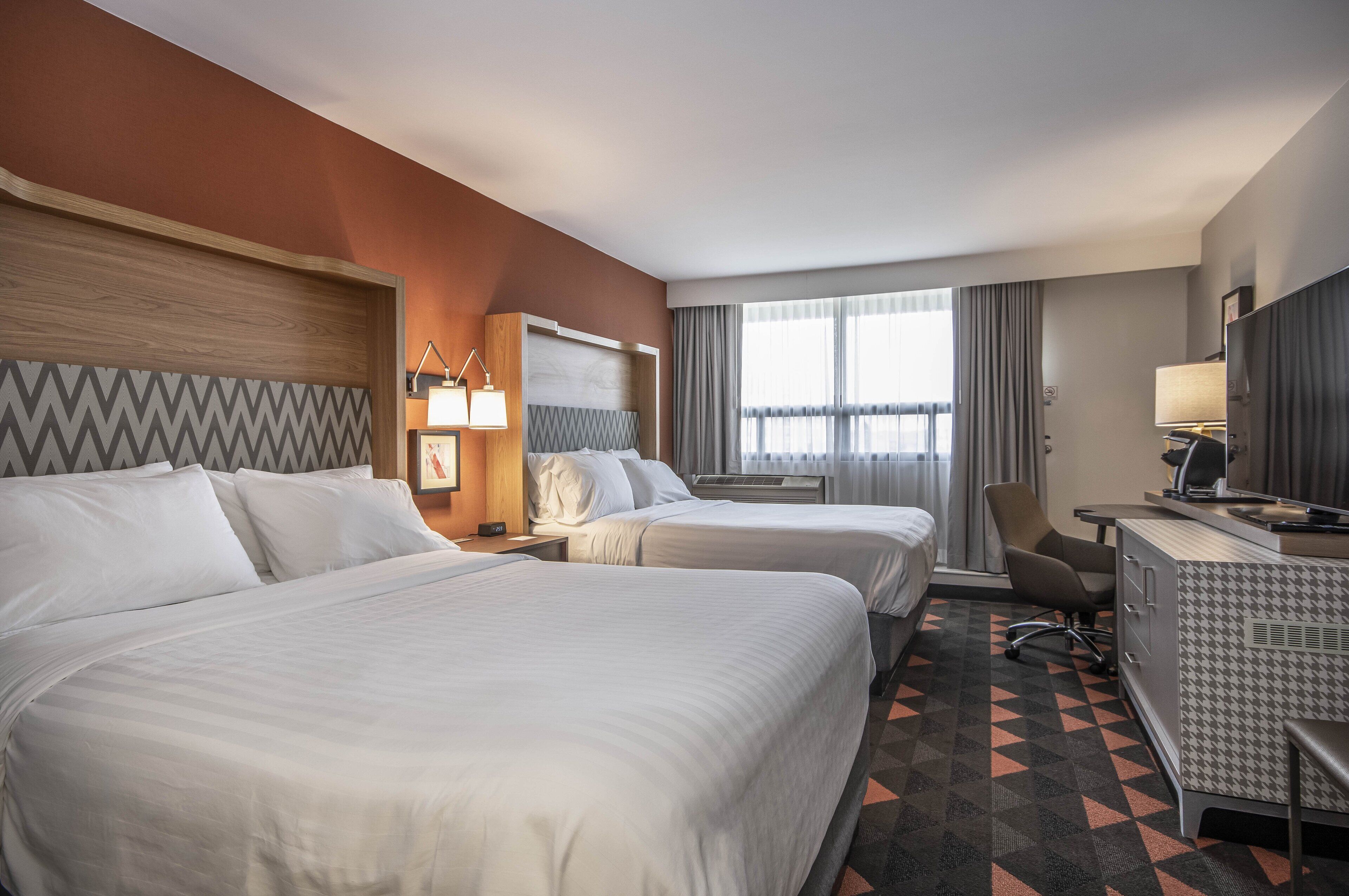 A hotel room with two beds, a desk, a chair, a TV, and a window with curtains.