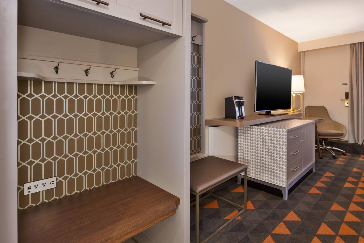 Une chambre d’hôtel avec un mur orné de motifs, un bureau avec un téléviseur à écran plat, une lampe, une chaise et une moquette à motifs.