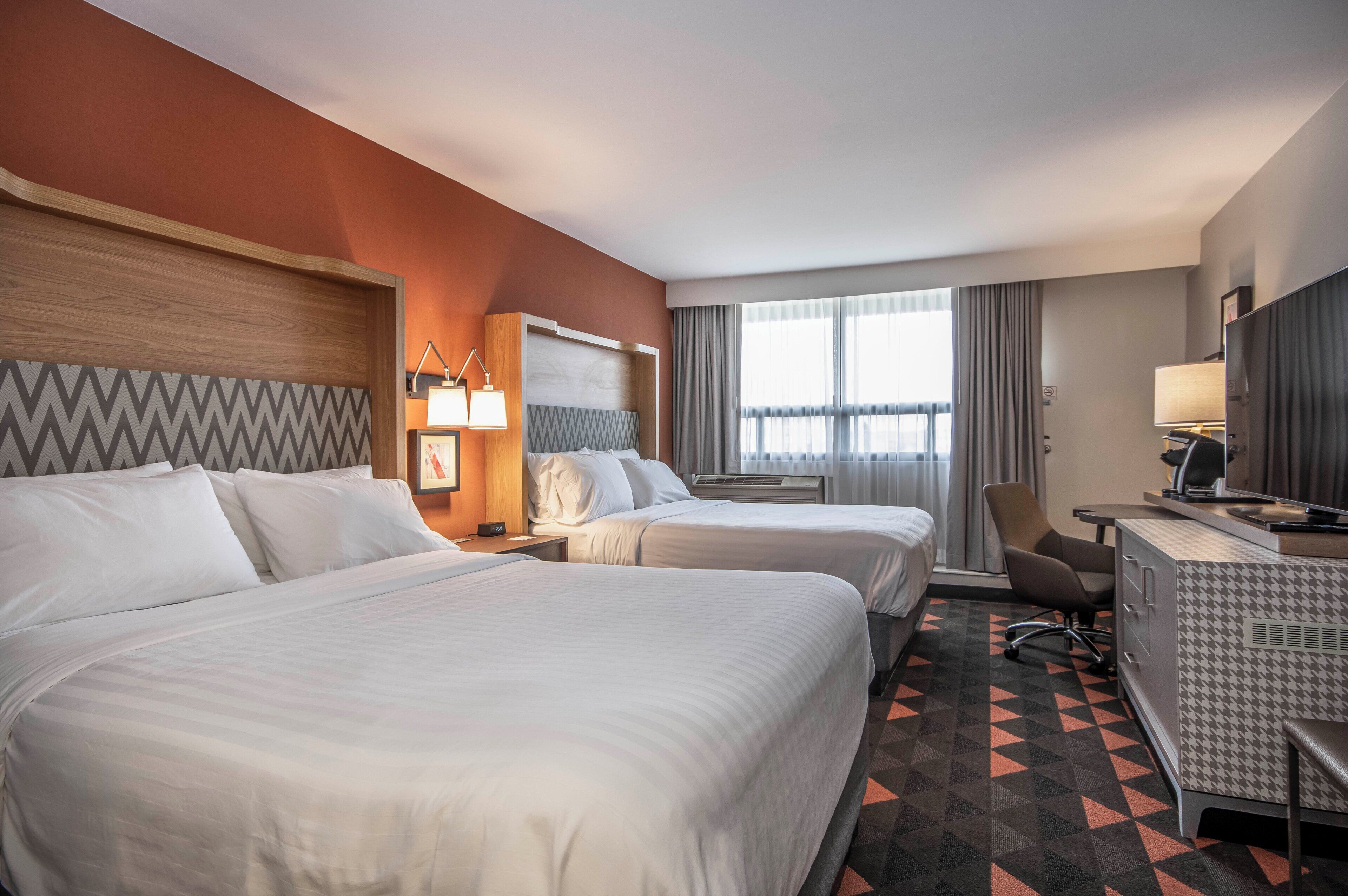 A hotel room with two beds, a desk, a chair, a lamp, and a window with curtains.