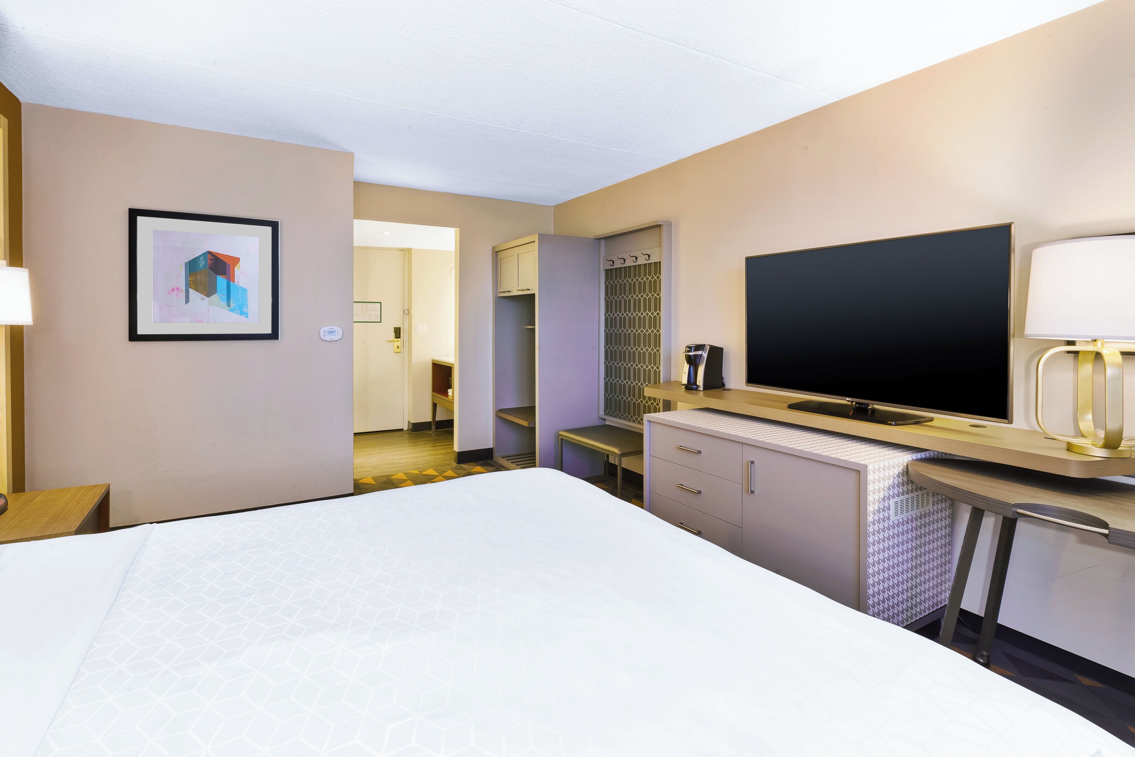 A hotel room with a bed, a flat-screen TV, a desk with a lamp, a mirror, and a framed picture on the wall.