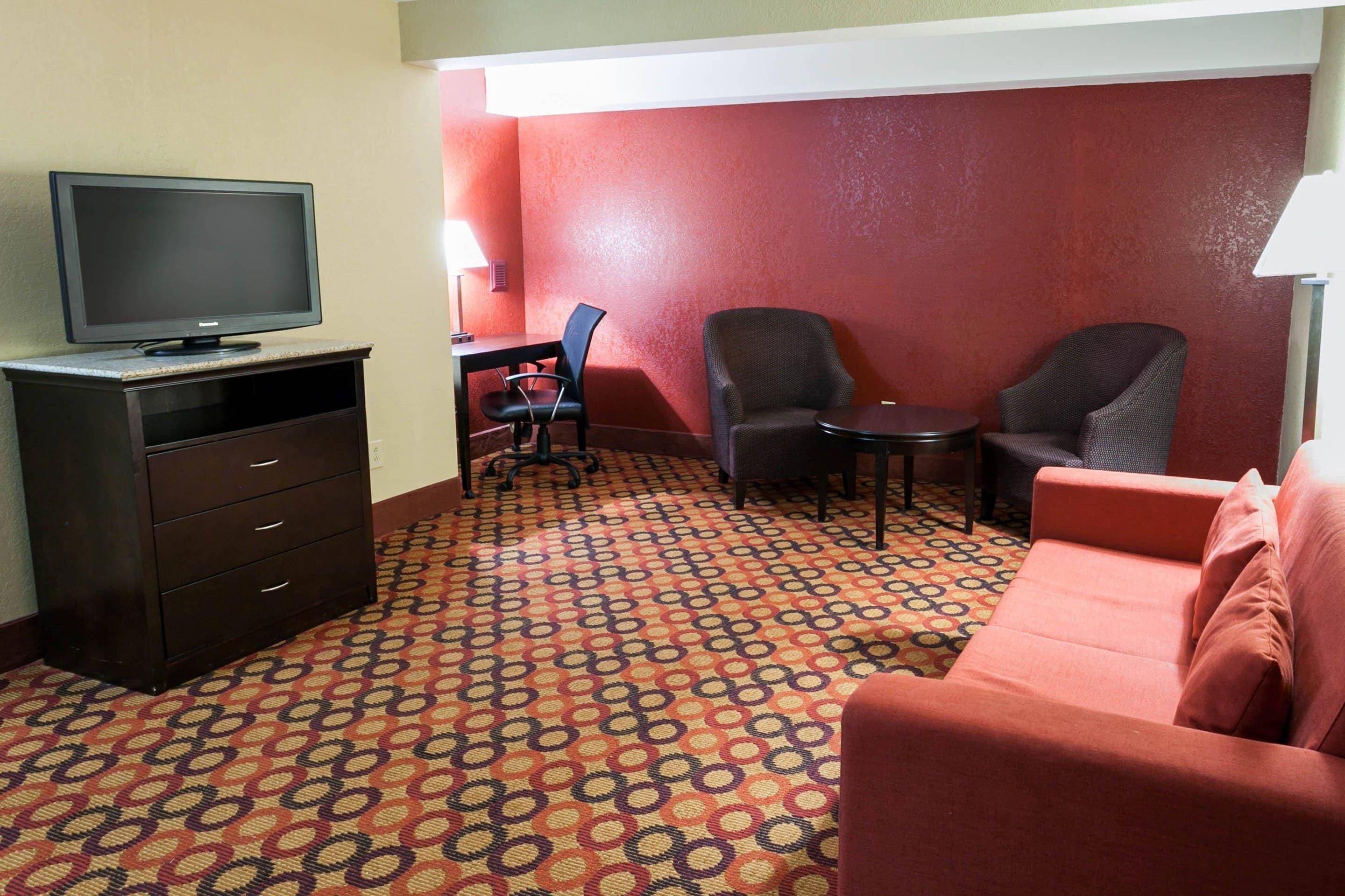 A hotel room with a flat-screen TV on a dresser, two chairs, a small table, a red sofa, and a patterned carpet.