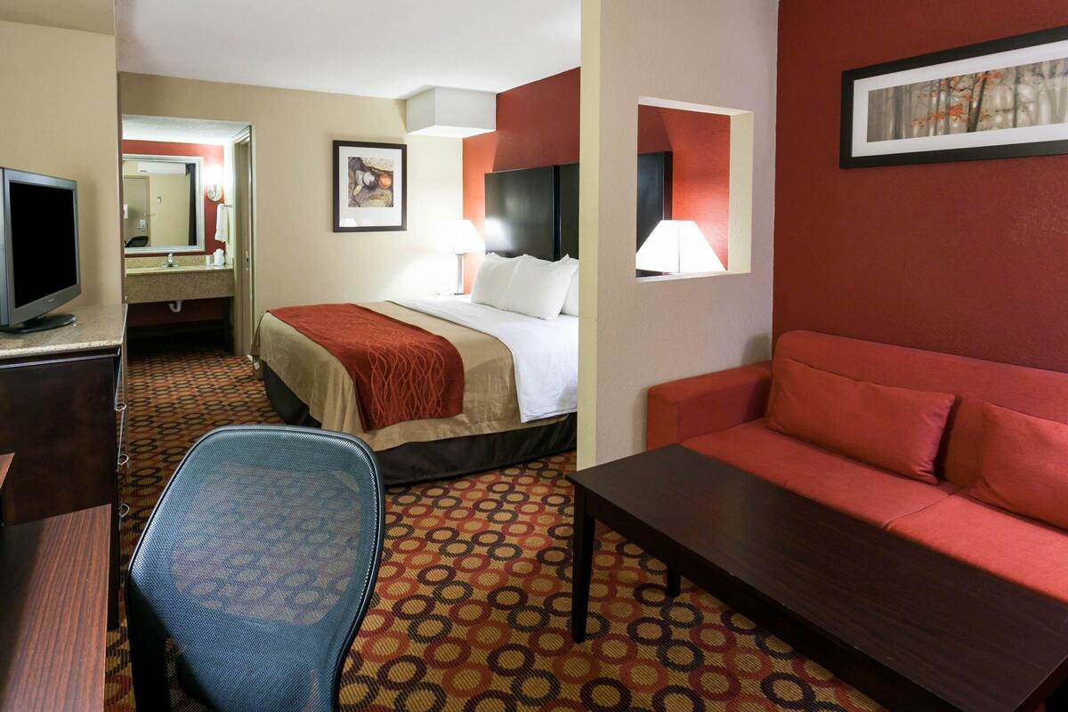 Ein Hotelzimmer mit einem Bett, einem Schreibtisch mit Computer, einem Sessel und einer Couch, sowie einem gemusterten Teppich.