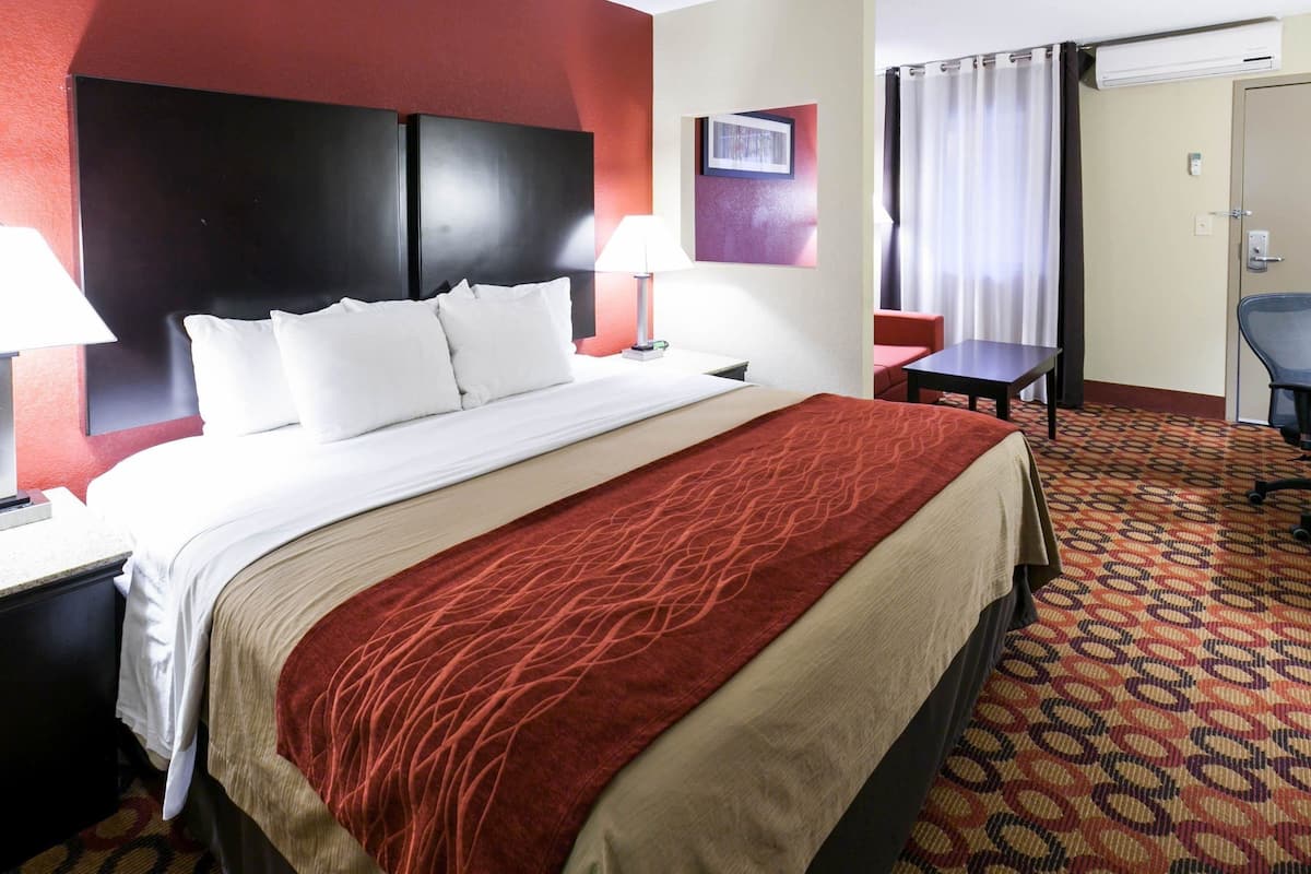 Ein Hotelzimmer mit einem großen Bett, zwei Nachttischen mit Lampen, einem kleinen Schreibtisch, einem Stuhl und einer Tür mit Vorhängen.