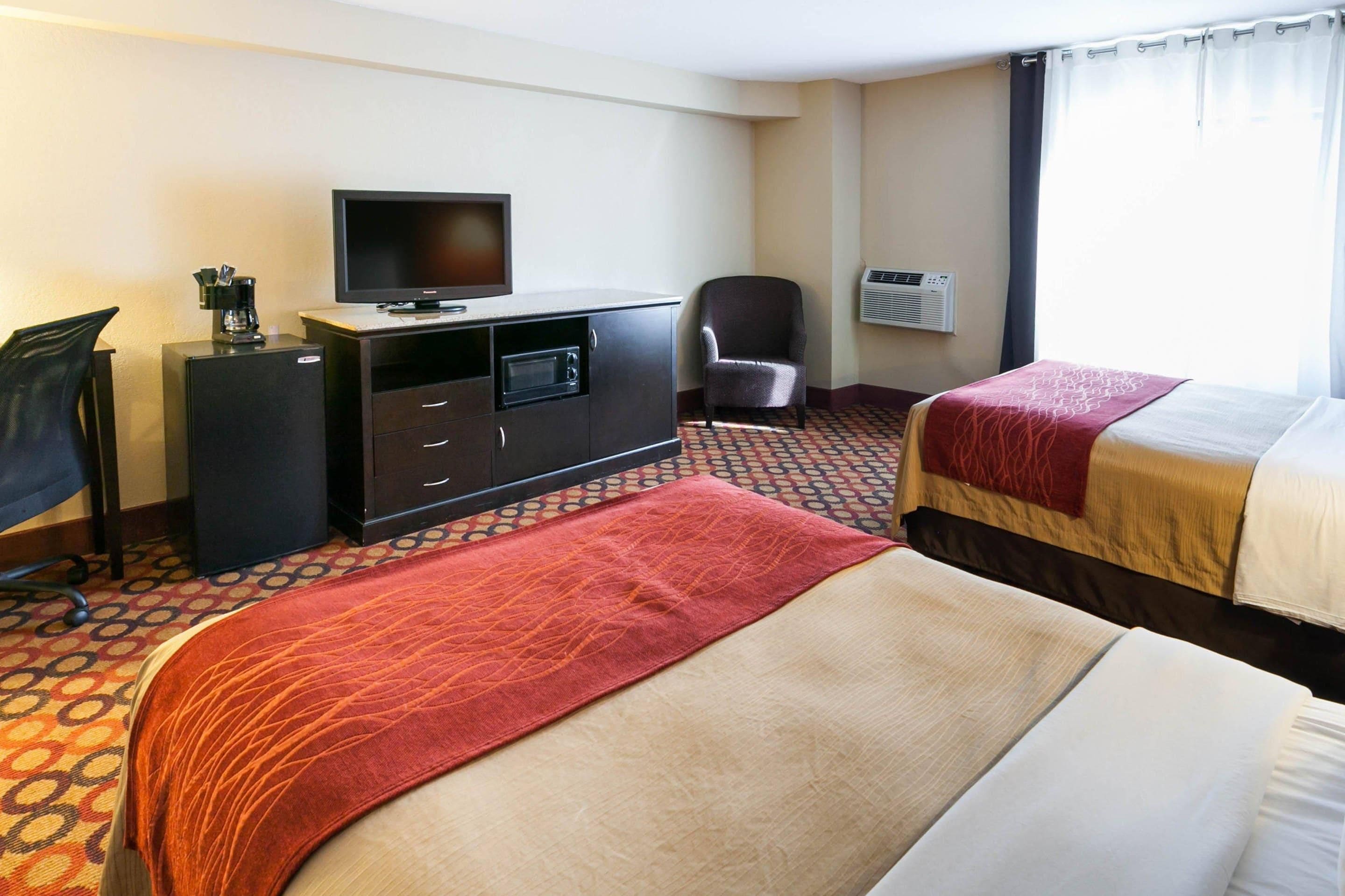 A hotel room with two beds, a desk with a television, a chair, and an air conditioning unit.