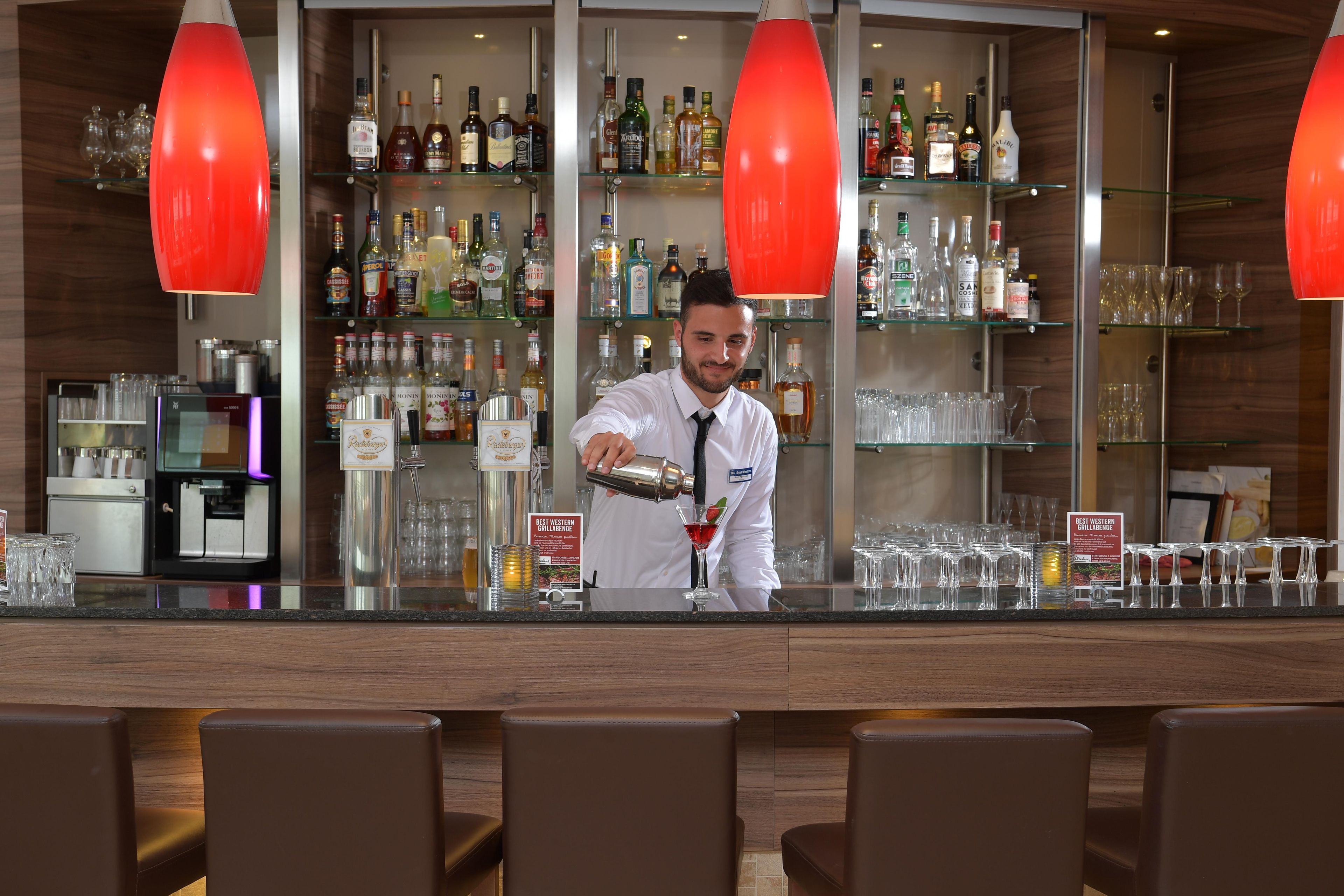 A bar area with a bartender preparing a drink, shelves of various liquor bottles, and a coffee machine.