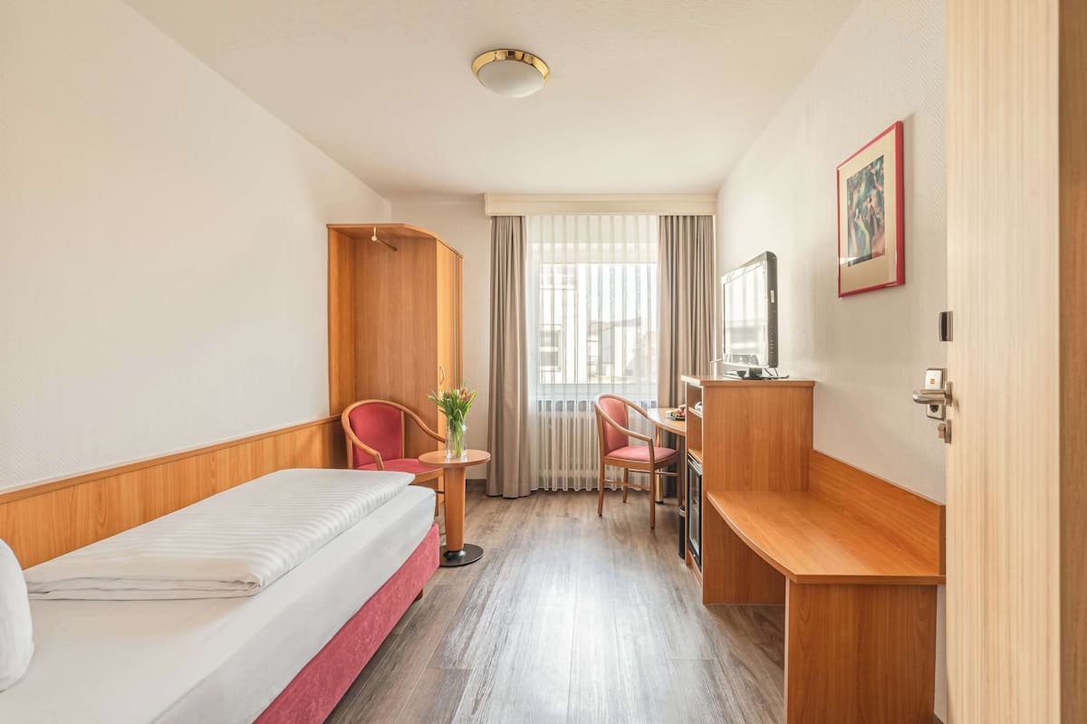 A hotel room with a bed, a desk, two chairs, a small table with a vase, and a window with curtains.