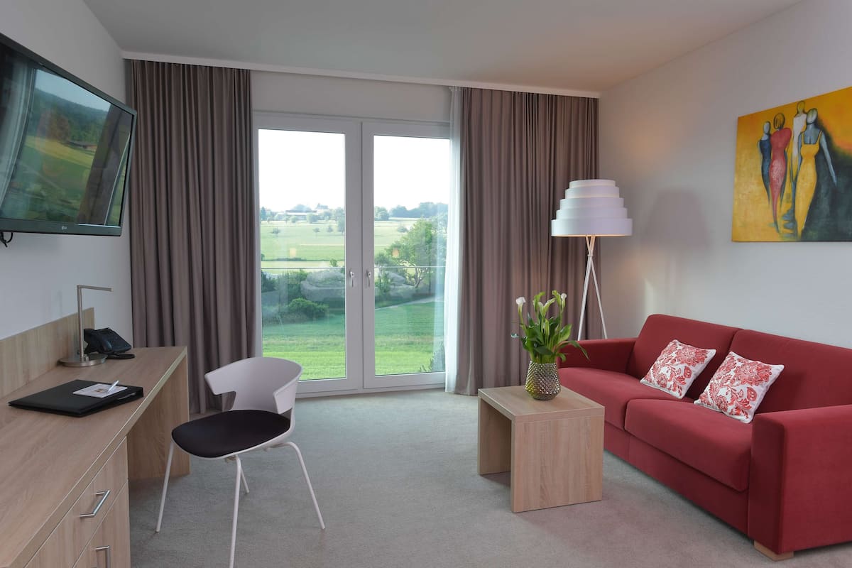 A modern hotel room with a desk, chair, sofa, and a large window with a view of the countryside.