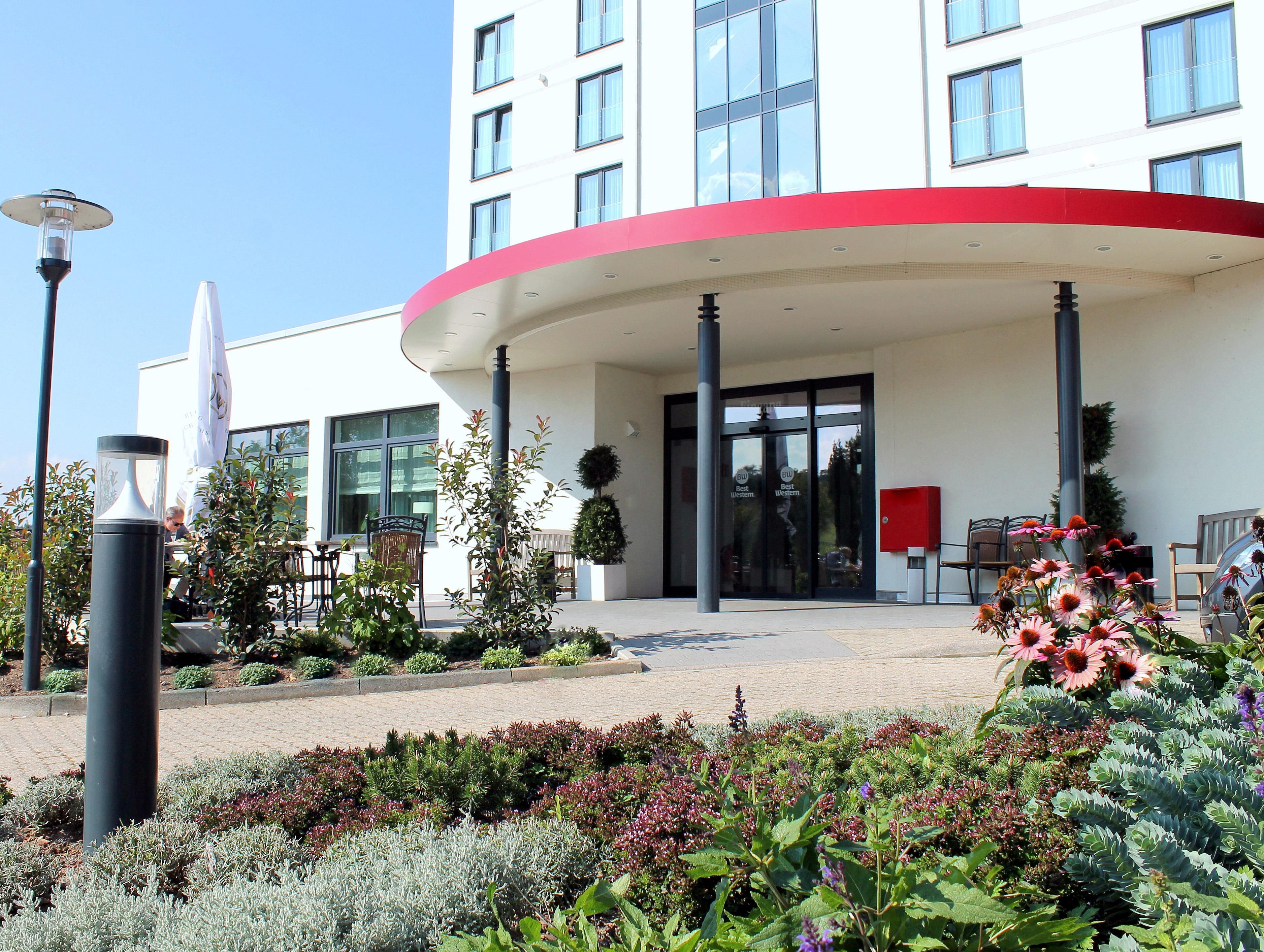 A hotel entrance with a curved facade, a glass door entrance, and a landscaped garden area with various plants and flowers.