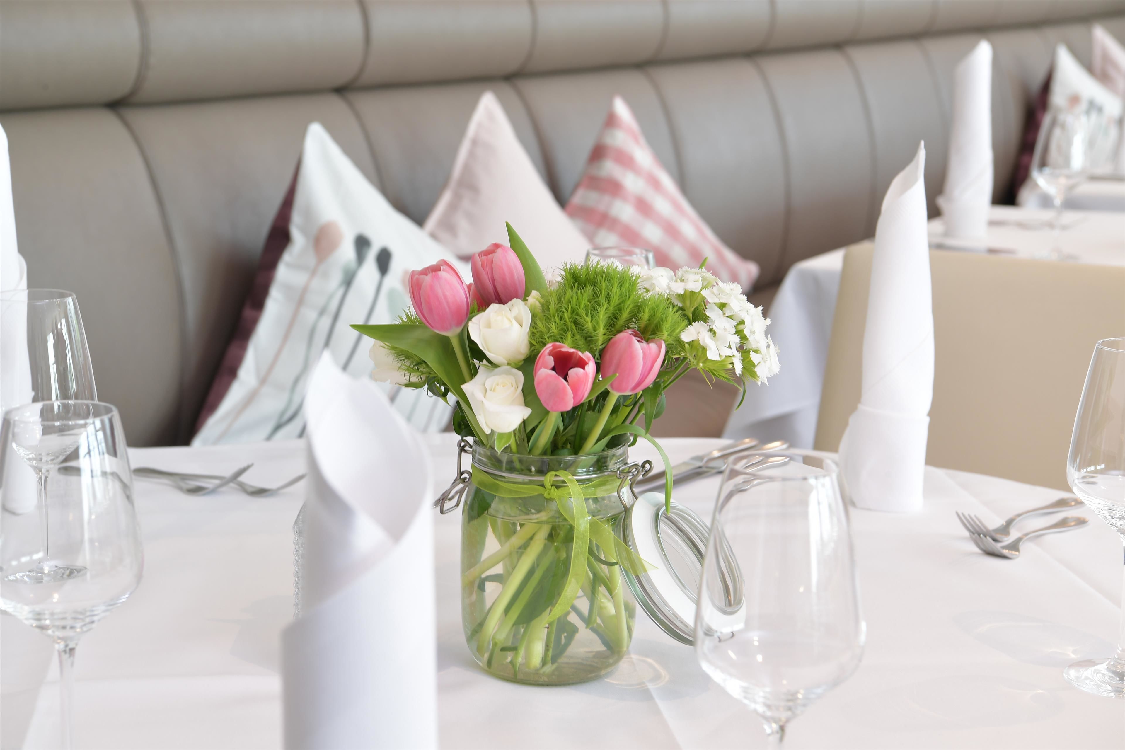 A dining table with a glass vase of flowers, white tablecloth, and neatly arranged cutlery.