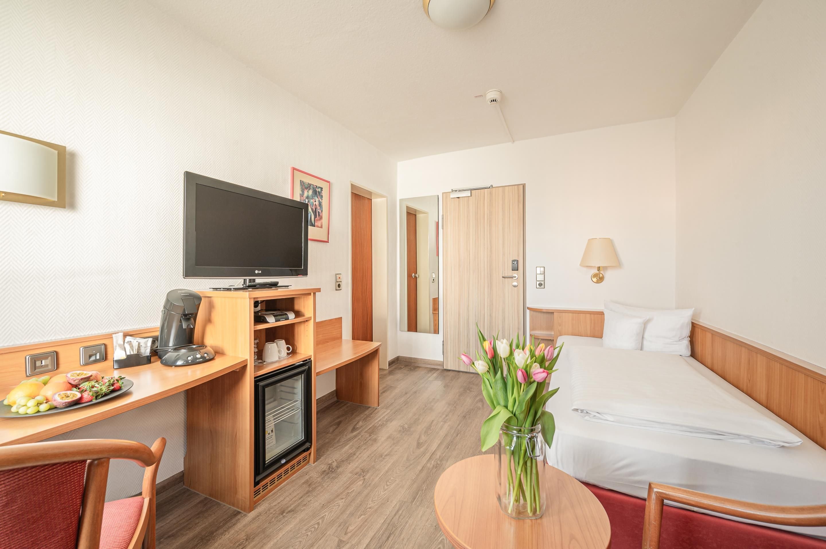 A hotel room with a bed, a desk with a television, a coffee machine, and a vase with flowers.
