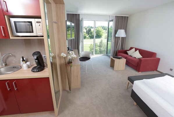 A hotel room with a kitchenette, a bed, a red sofa, a small table, and a view of the outdoors.