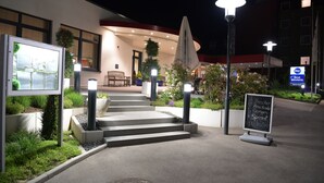 Entrada de hotel con escalones que conducen a una zona cubierta, mobiliario exterior y un cartel.