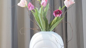 Eine weiße Vase mit rosa und violetten Tulpen auf einer Holztischplatte vor grauem Vorhang.