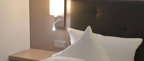 Una cama bien hecha con ropa de cama blanca, cabecera, mesita de noche y lámpara fijada a la pared.