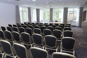 Un salón de conferencias con filas de sillas negras, una mesa con proyector y ventanas grandes con cortinas.