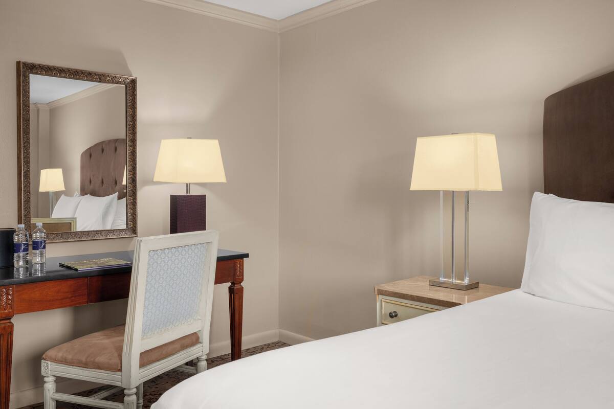 Une chambre d’hôtel comprenant un lit, un bureau avec une chaise, une table de chevet avec une lampe et un miroir au mur.
