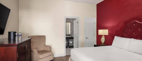 Una habitación de hotel con cama, una silla, un escritorio, televisor y puerta que da a un baño.