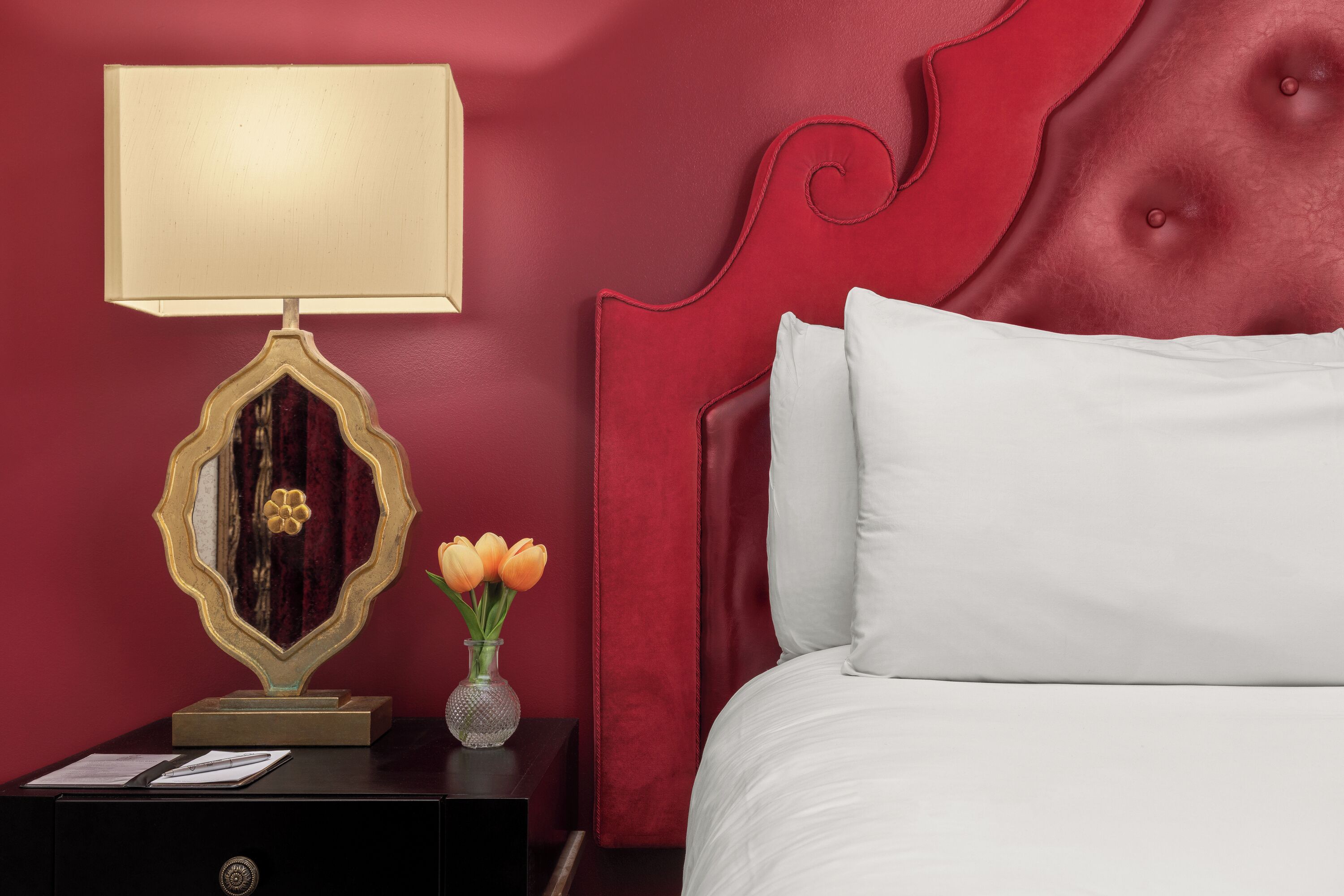 A bedroom with a red headboard, a bedside table with a lamp, a vase with flowers, and a bed with white bedding.