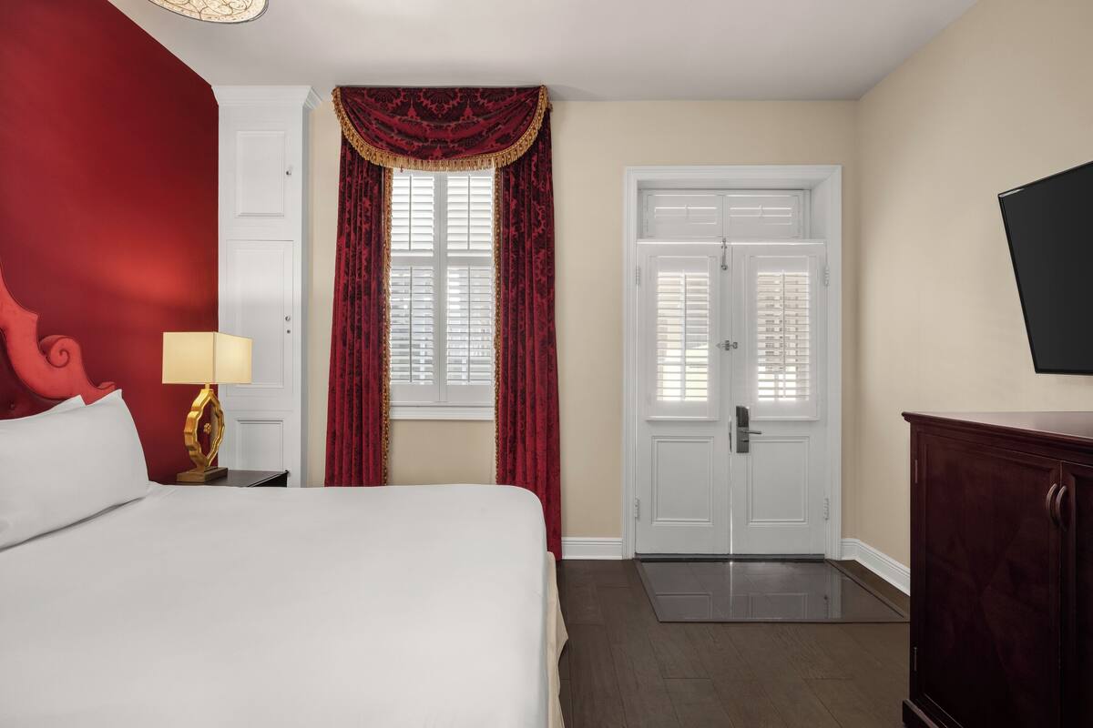 Une chambre d’hôtel avec un lit, une table de chevet avec une lampe, une armoire, une porte avec un cadre blanc et une fenêtre avec des rideaux rouges.