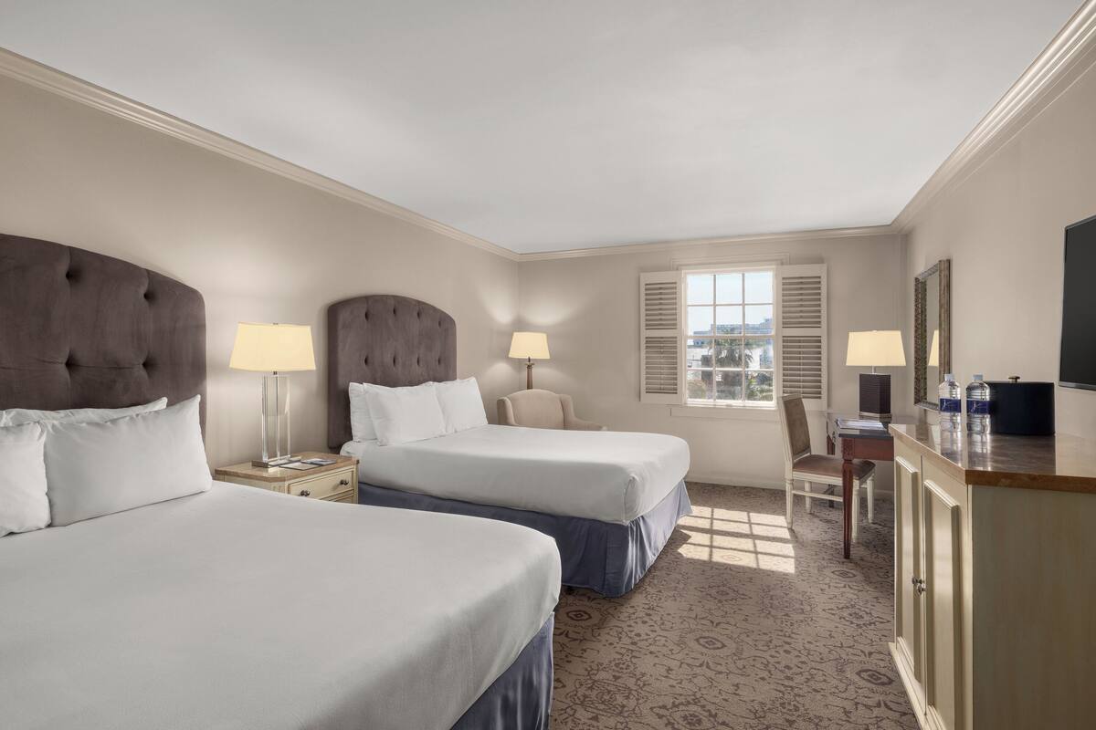 Une chambre d’hôtel avec deux lits, un bureau, une chaise, une télévision et une fenêtre avec des volets.