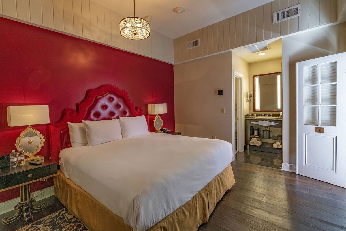 Une chambre d’hôtel avec un grand lit, un mur d’accent rouge, un lustre, une table de chevet et une salle de bain visible à travers une porte ouverte.