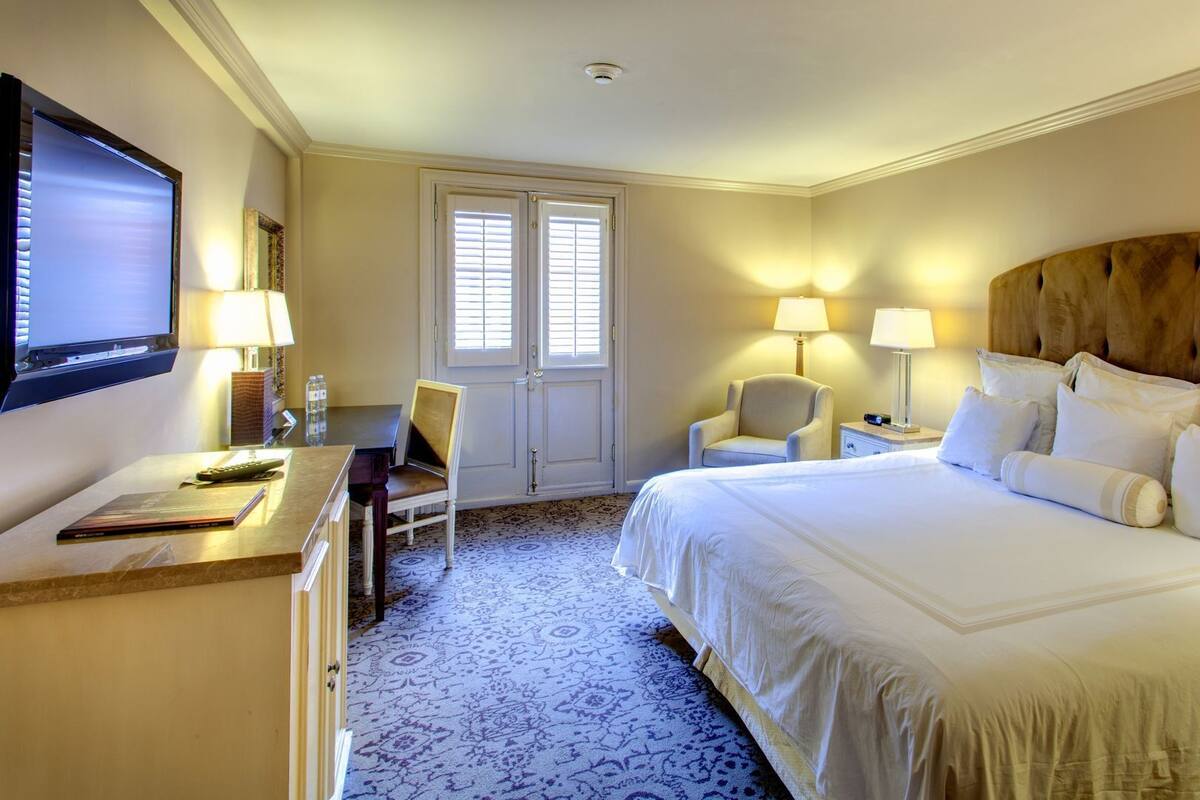 Une chambre d’hôtel comprenant un lit, deux lampes de chevet, un bureau avec une télévision, une chaise et une porte avec des fenêtres.