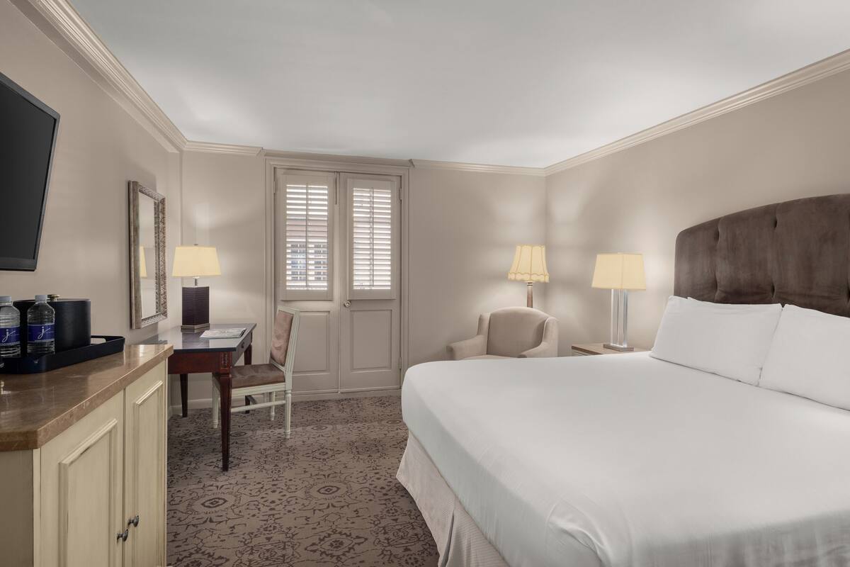 Une chambre d’hôtel équipée d’un lit, d’un bureau, d’une chaise, d’une télévision et d’une fenêtre avec des stores.