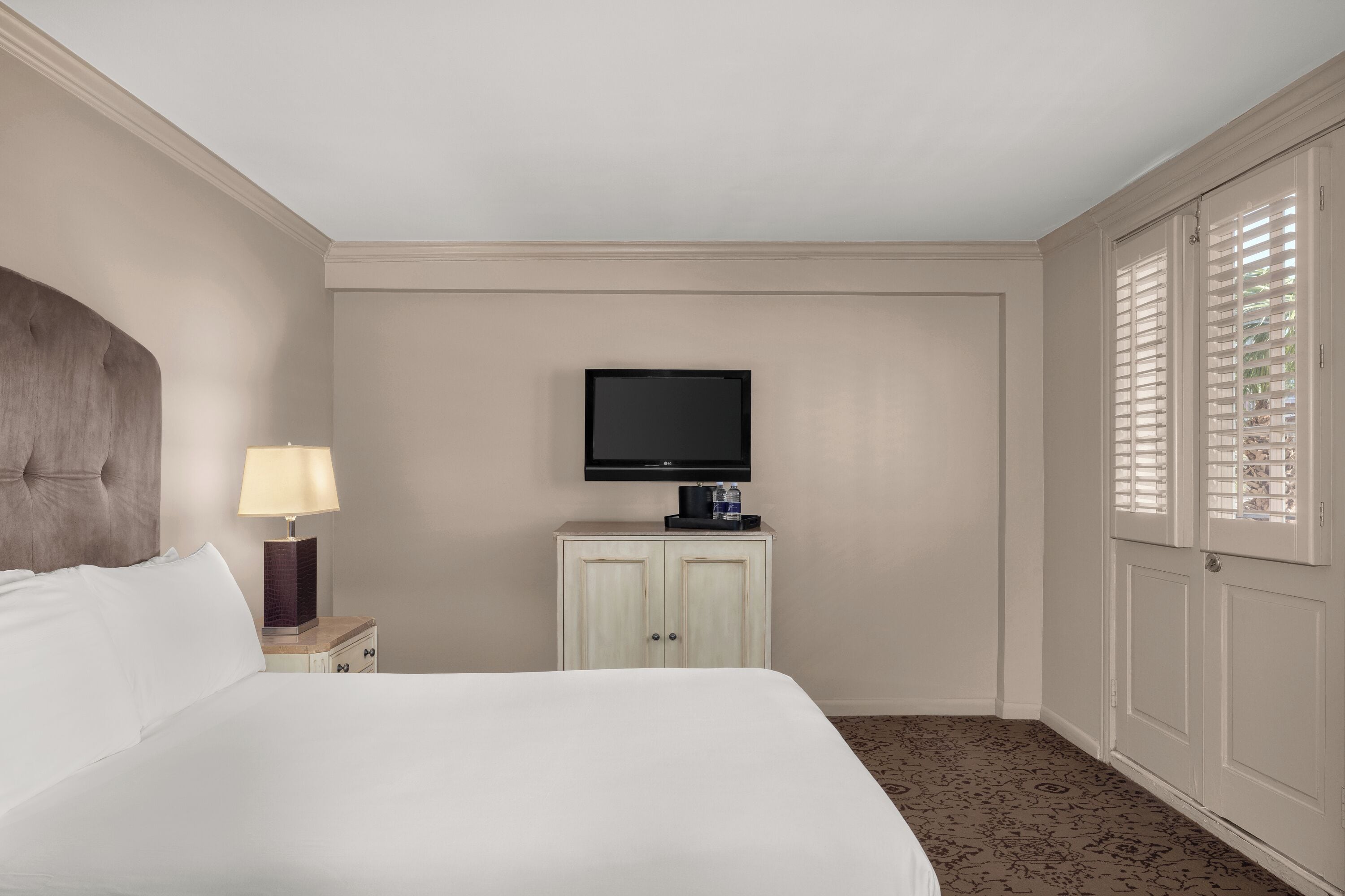 Una habitación de hotel con una cama grande, un televisor en un mueble, una mesita de noche con lámpara y una ventana con persianas.