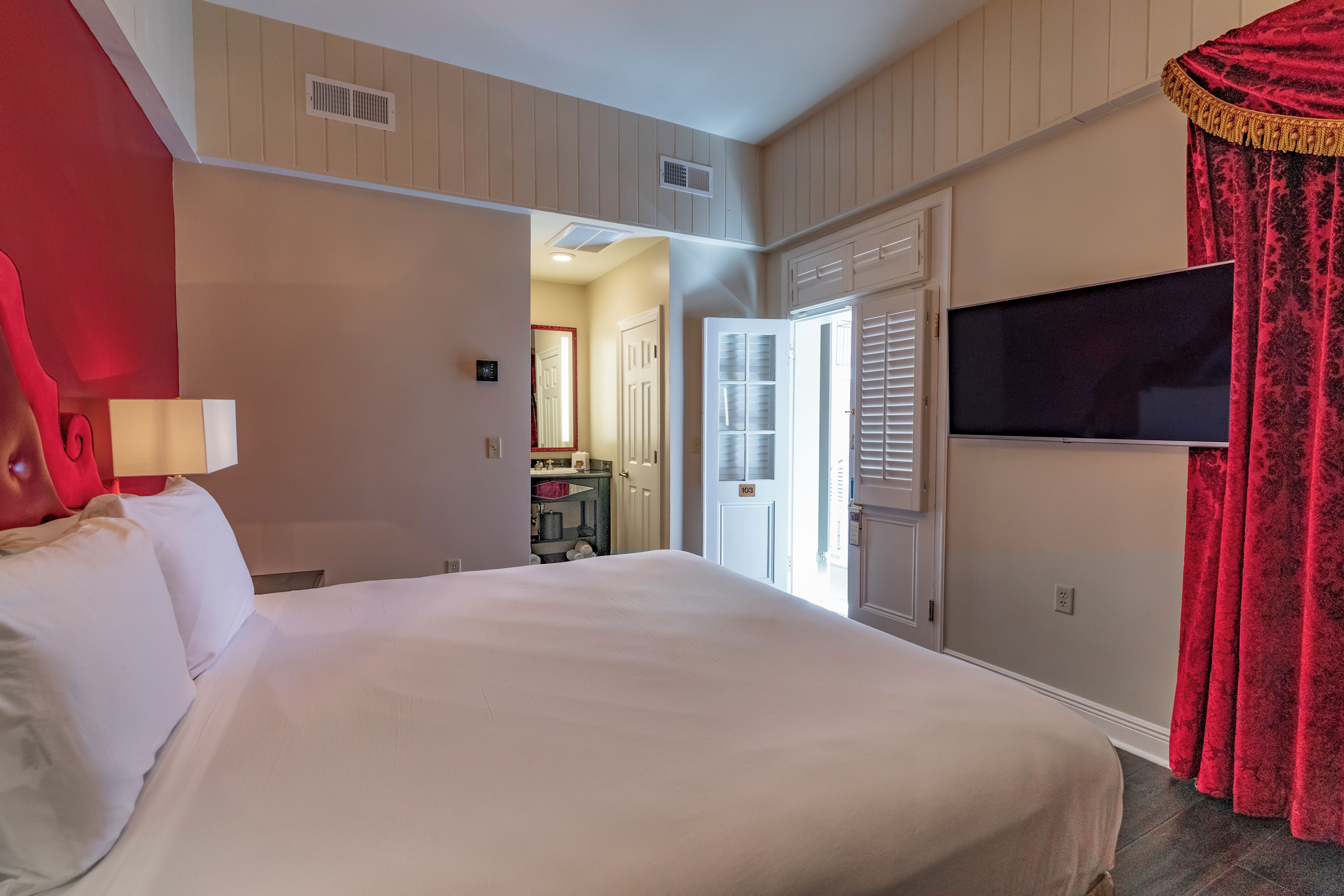 A hotel room with a bed, a television, a red curtain, and a door leading to a bathroom.