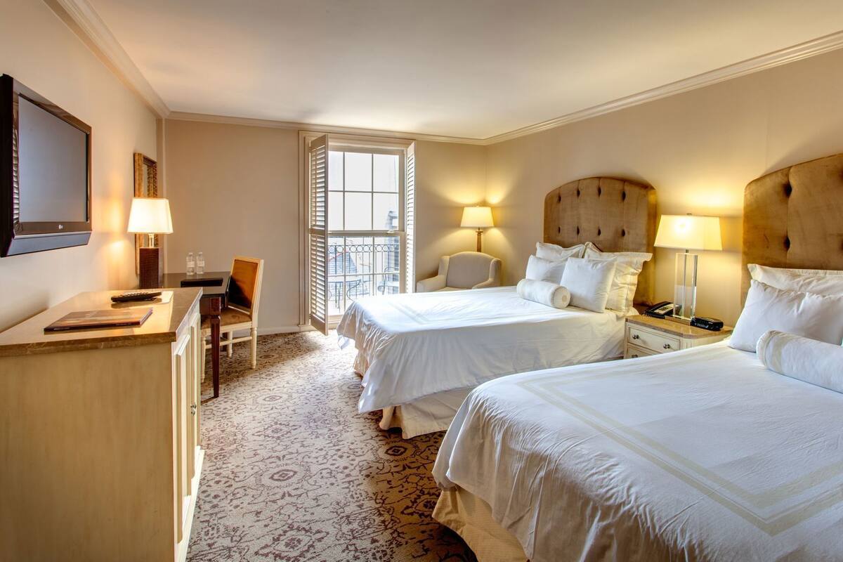 Une chambre d’hôtel avec deux lits, un bureau, une chaise, une télévision et une fenêtre avec des stores.