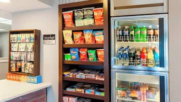 Mostrador de snacks y bebidas en el vestíbulo de un hotel, con estantes que incluyen Doritos y Ruffles, y un refrigerador lleno de bebidas como Mountain Dew y Gatorade.