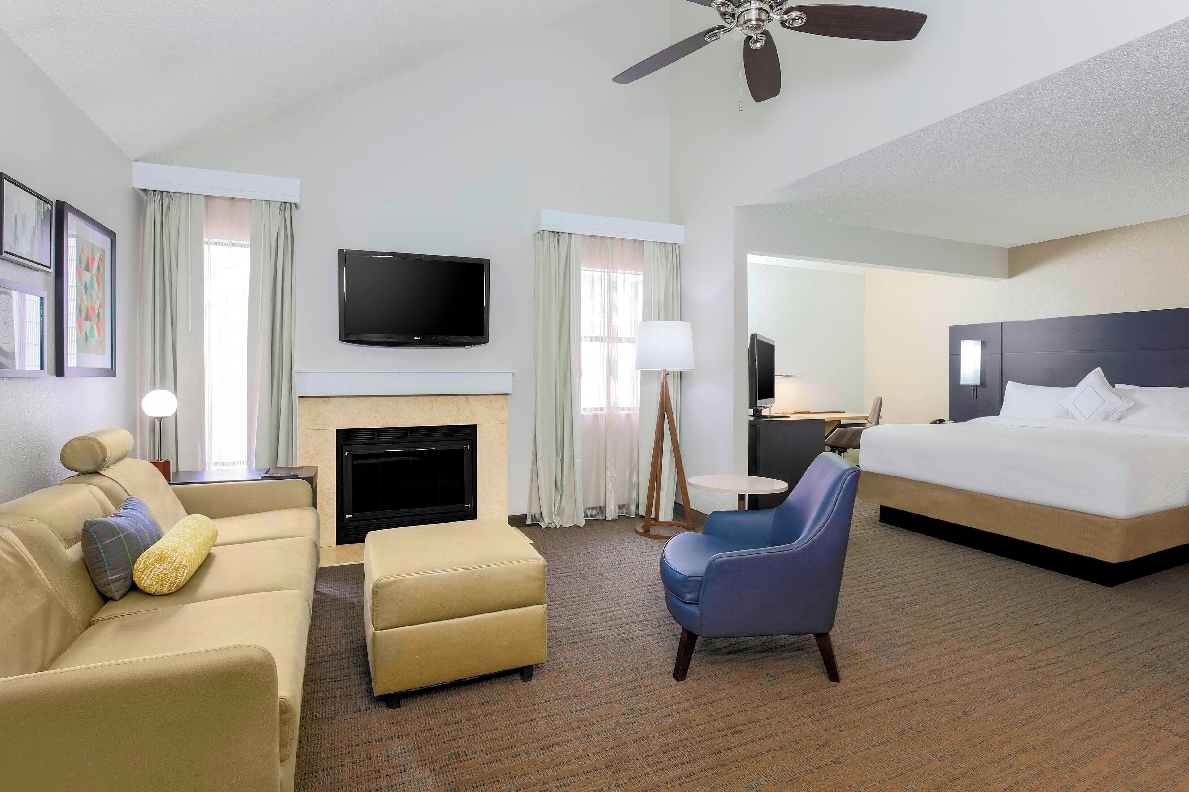 Une chambre d’hôtel équipée d’un canapé, d’un pouf, d’un fauteuil, d’un lit, d’une télévision à écran plat, d’une cheminée, d’un ventilateur de plafond et d’un grand miroir.